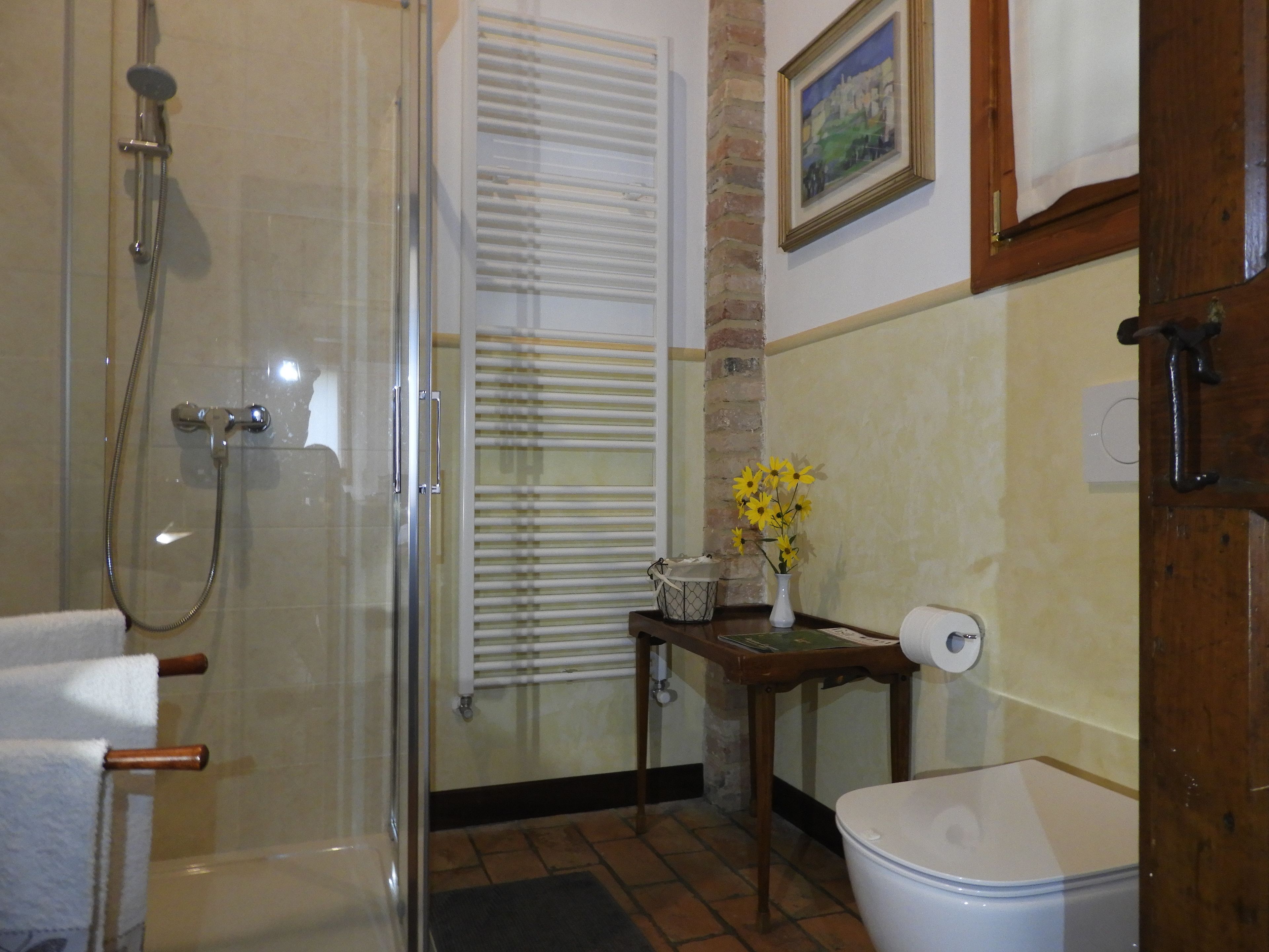 A bathroom with a glass shower enclosure, a toilet, a small table with a vase of flowers, and a window with blinds.
