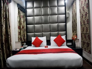 A hotel room with a large bed, a tufted headboard, patterned curtains, and a nightstand with a vase of flowers. - OYO 464 Hotel Lotus Palace (New Delhi)