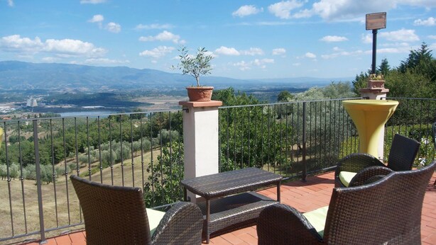 Una terraza con mobiliario de mimbre, una planta en maceta y vistas a un paisaje que incluye una masa de agua y montañas.