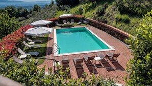 A rectangular swimming pool with a stone border, surrounded by lounge chairs and umbrellas, set against a backdrop of hills and trees. - B&B Amoliv (Cavriglia)