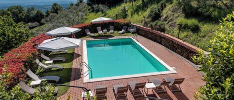 A rectangular swimming pool with a stone border, surrounded by lounge chairs and umbrellas, set against a backdrop of hills and trees.