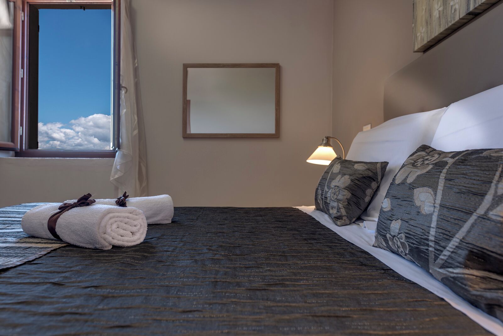 Ein ordentlich gemachtes Bett mit dunklem Kopfteil, einer Nachttischlampe und einem Fenster mit Blick auf Wolken.