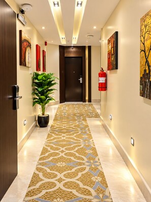 A hotel corridor with a patterned carpet, wall-mounted artwork, a potted plant, a fire extinguisher, and a door with a keypad lock.