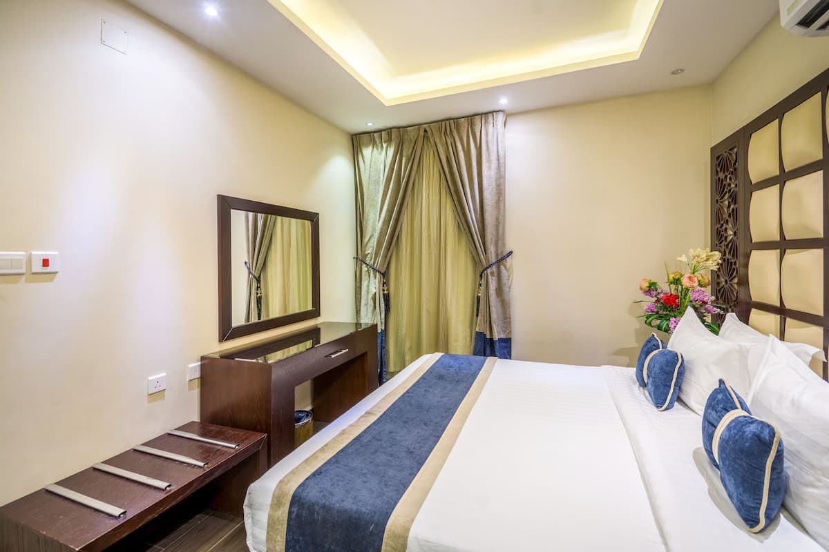 Una habitación de hotel con cama, escritorio, espejo, ventana con cortinas y un panel decorativo.
