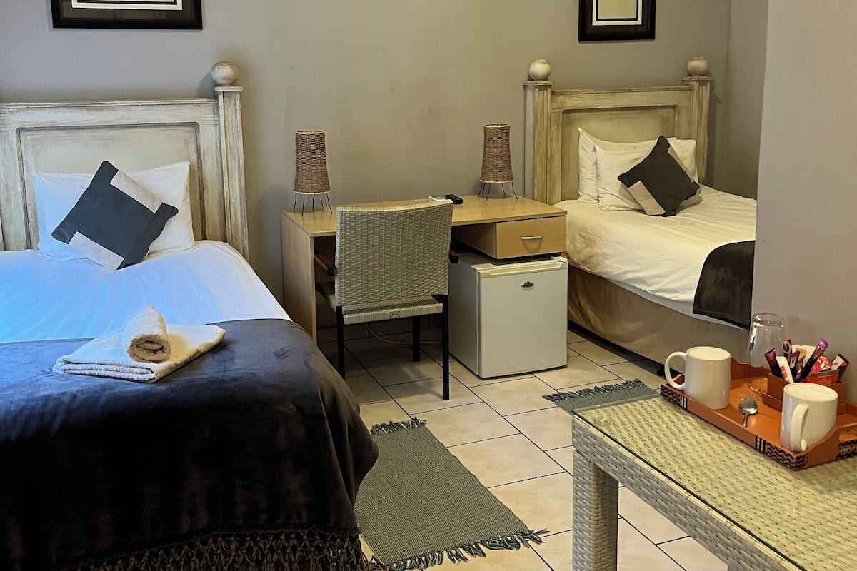 A hotel room with two beds, a small table with mugs and snacks, a chair, and framed pictures on the walls.