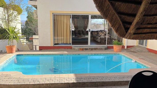 Una piscina con techo de paja, un patio con plantas en macetas y vista a un edificio moderno con puertas de vidrio correderas.