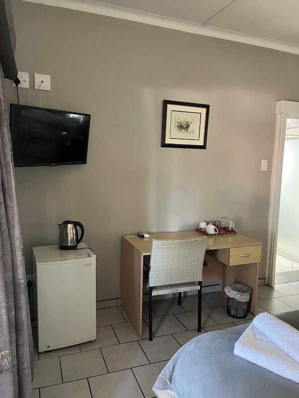 Habitación de hotel con una cama, un escritorio con una silla, un televisor, un refrigerador pequeño, un hervidor de agua y un cuadro en la pared.