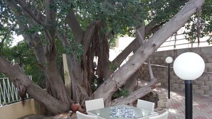 Un área de estar al aire libre con una mesa de vidrio y sillas de mimbre blanco, rodeada de árboles grandes y un muro de ladrillo.
