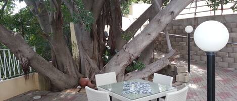 An outdoor seating area with a glass-top table and white wicker chairs, surrounded by large trees and a brick wall.