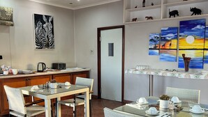 Un comedor con una mesa de vidrio y sillas de mimbre, una zona de cocina con cafetera, y decoración en la pared que muestra cebras y una puesta de sol.