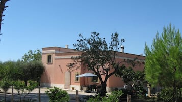Un edificio con tetto piatto, una finestra e una porta. Si vedono un tavolo e delle sedie all'esterno, e un albero di fronte all'edificio.