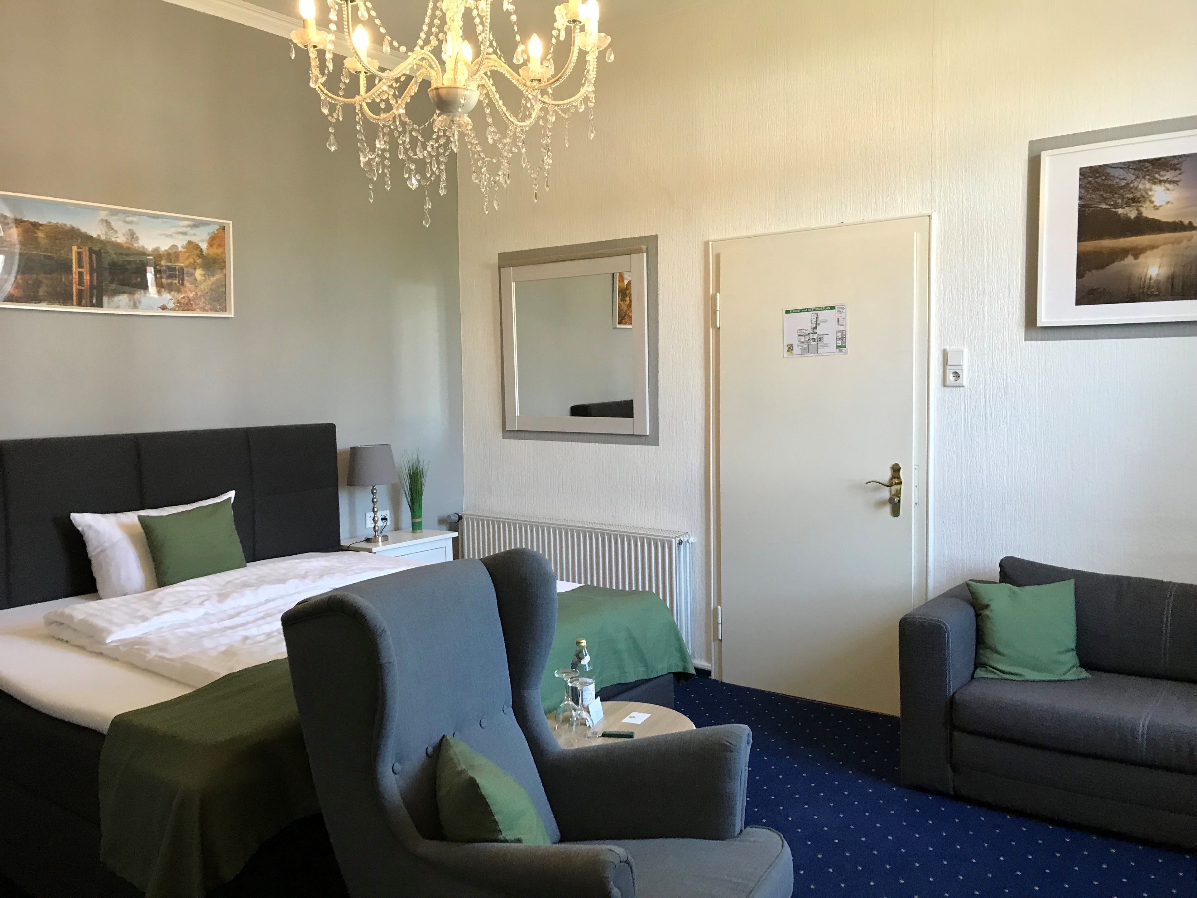 A hotel room with a bed, a chair, a sofa, a chandelier, a mirror, and a painting on the wall.