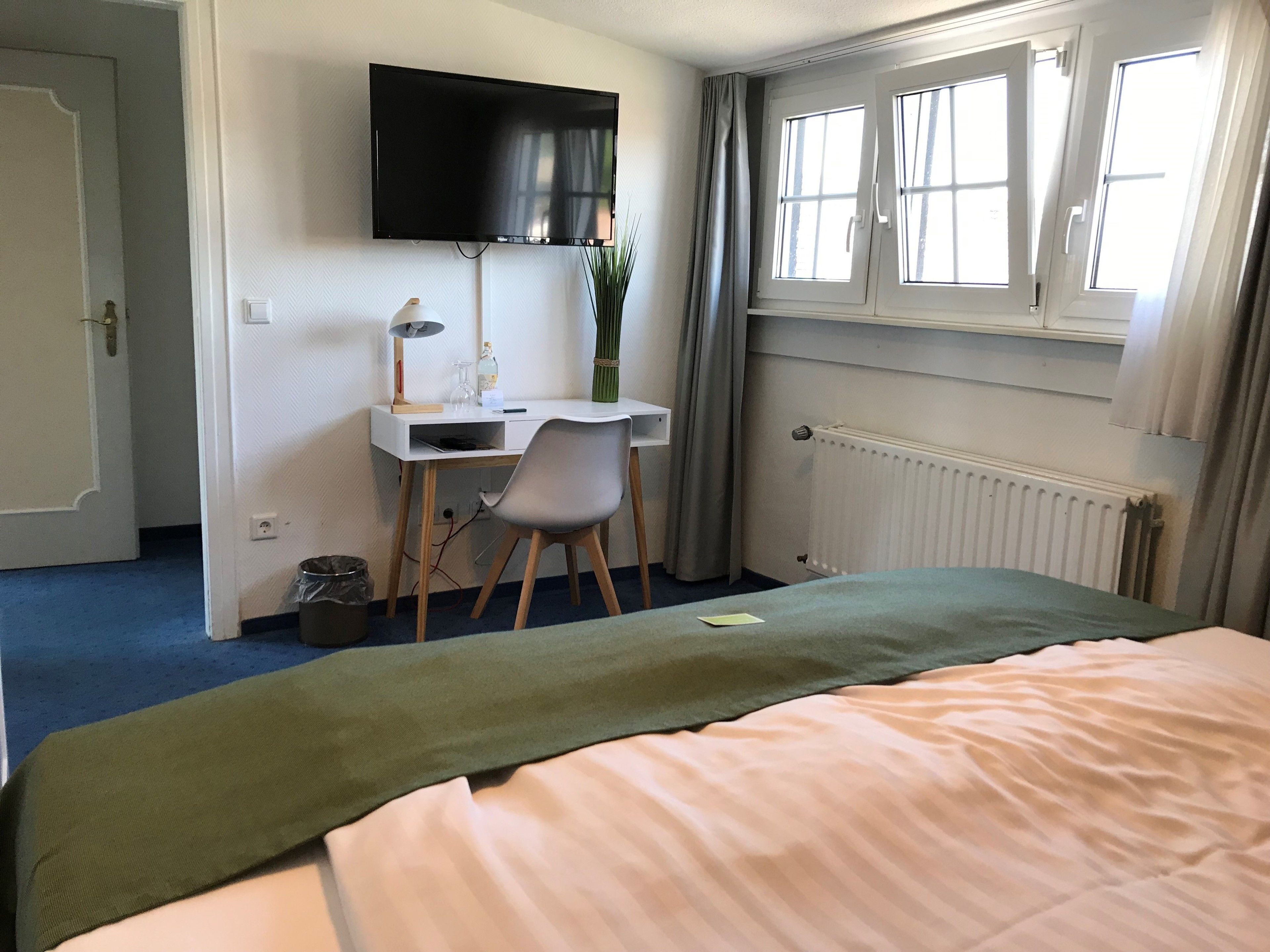 A bedroom with a bed, a desk, a chair, a TV, and a window with curtains.