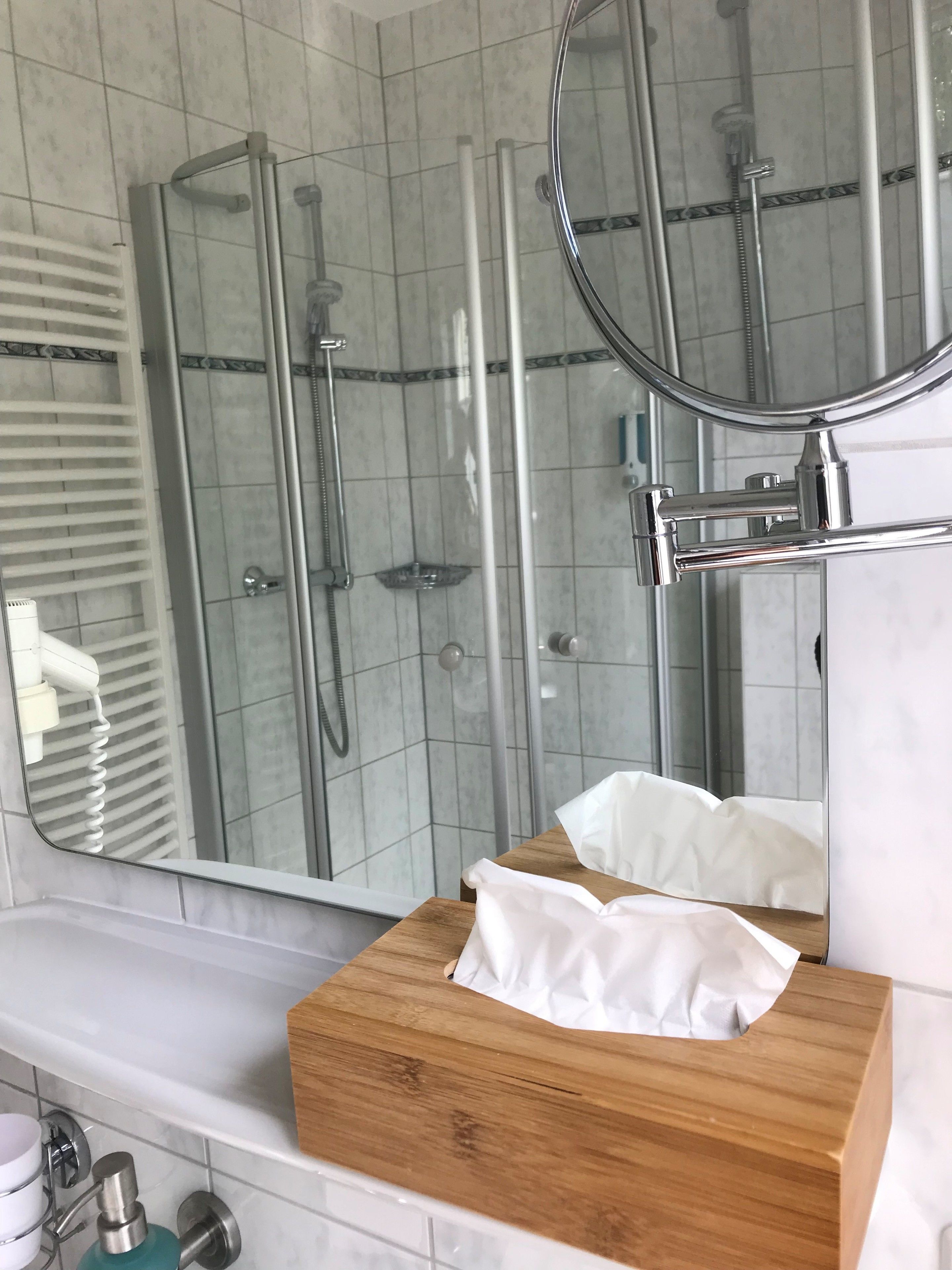 A bathroom with a shower, a mirror, a wooden vanity, and a towel dispenser.