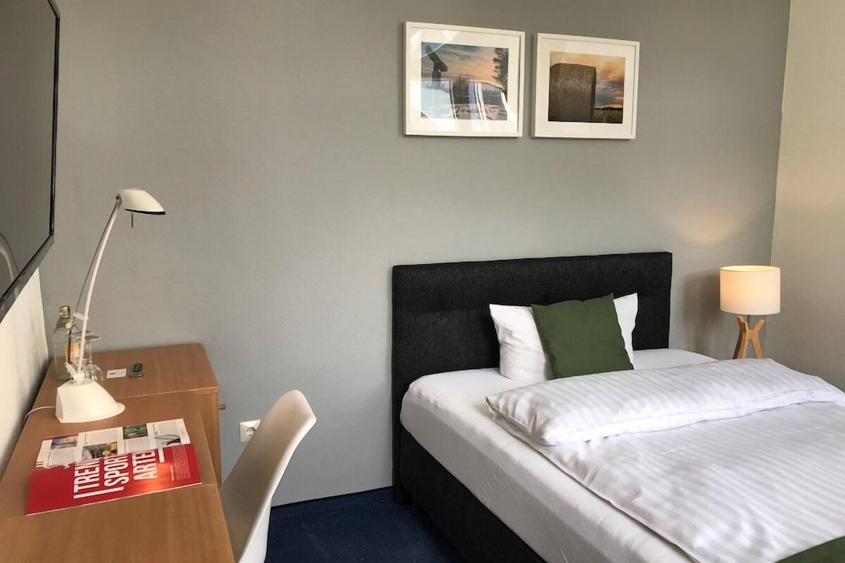 Quarto de hotel com uma cama, uma escrivaninha, uma cadeira, duas fotos emolduradas na parede e um abajur.