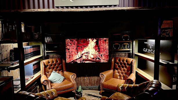 A cozy lounge area with leather armchairs, a television displaying a fireplace, and bookshelves filled with books.