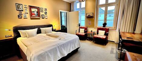 A hotel room with a large bed, two armchairs, a small table, and a window with curtains.