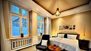 A hotel room with a large bed, a chair, a small table, and a window with a view of trees.