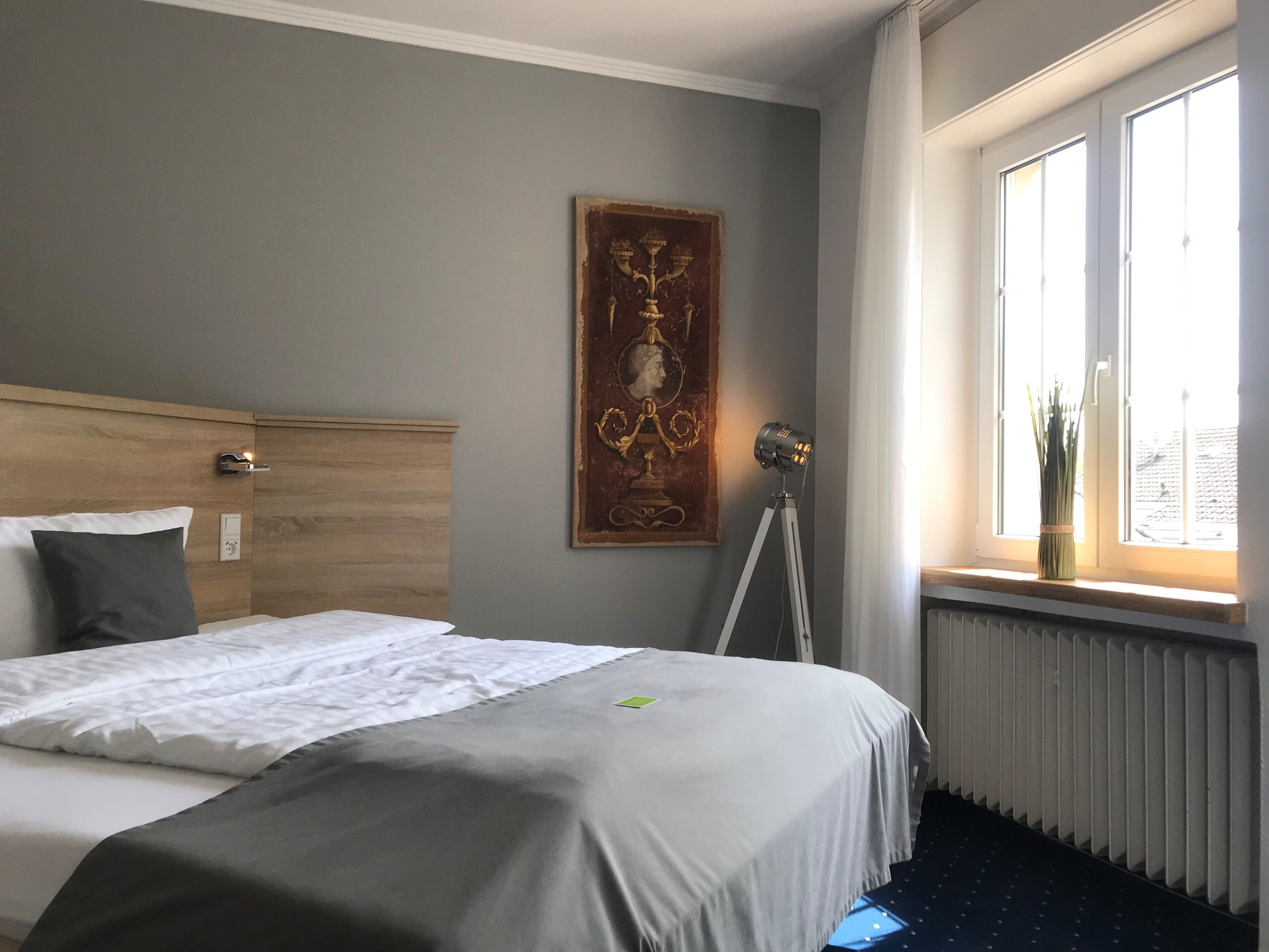 A hotel room with a bed, a painting on the wall, a window with a view, and a radiator.