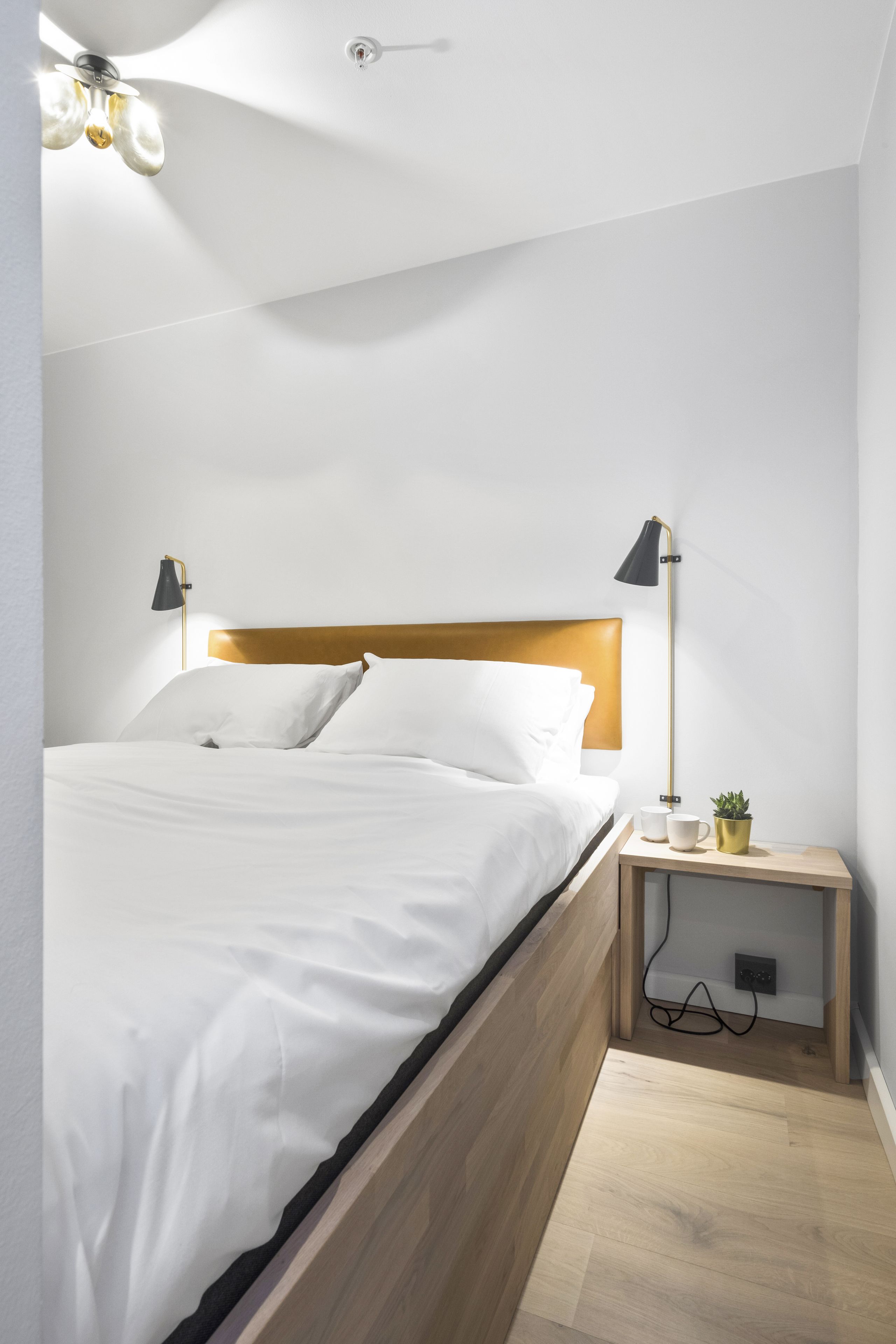 Un dormitorio moderno con cabecera de madera, cama con ropa blanca, dos lámparas de noche, una mesita de noche con una planta y un enchufe.