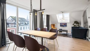 A modern living room with a wooden dining table, dark chairs, a sofa, and a large window offering a city view. - VANDER - Sea Story (Stavanger)