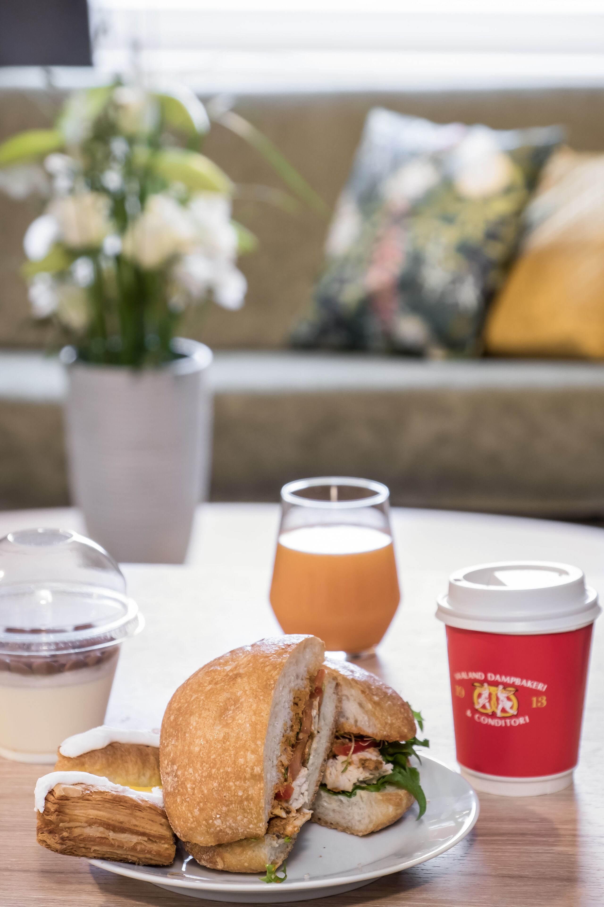 A sandwich on a plate, a glass of orange juice, a cup of coffee, and a vase with flowers on a table.