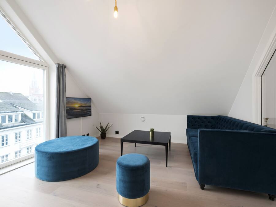 Un moderno loft con un'ampia finestra a lucernario, un divano in velluto blu scuro, due poggiapiedi rotondi, un tavolino nero e una pianta in vaso.
