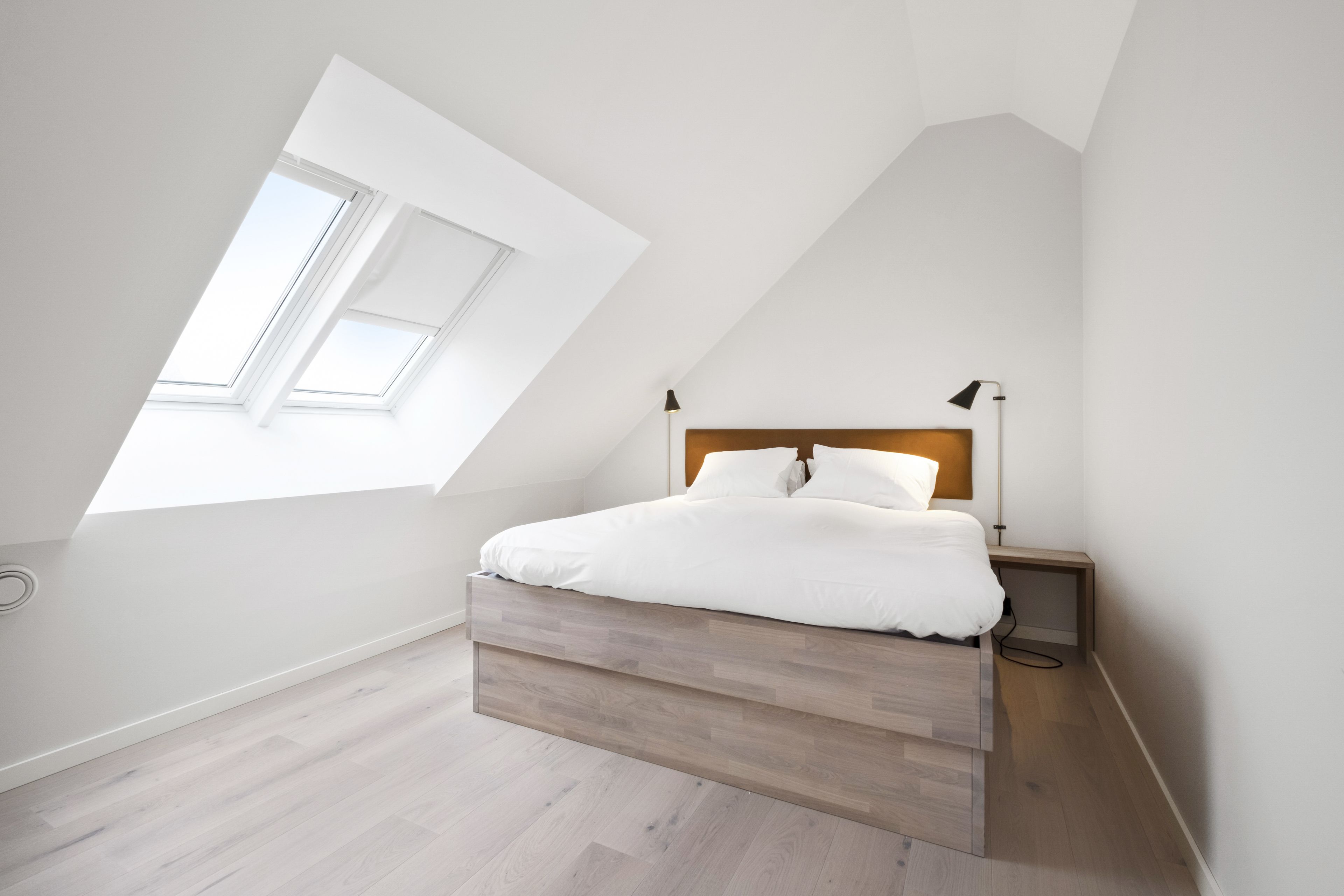 Quarto com teto inclinado, cama com roupa de cama branca, duas luminárias de cabeceira e um claraboia.