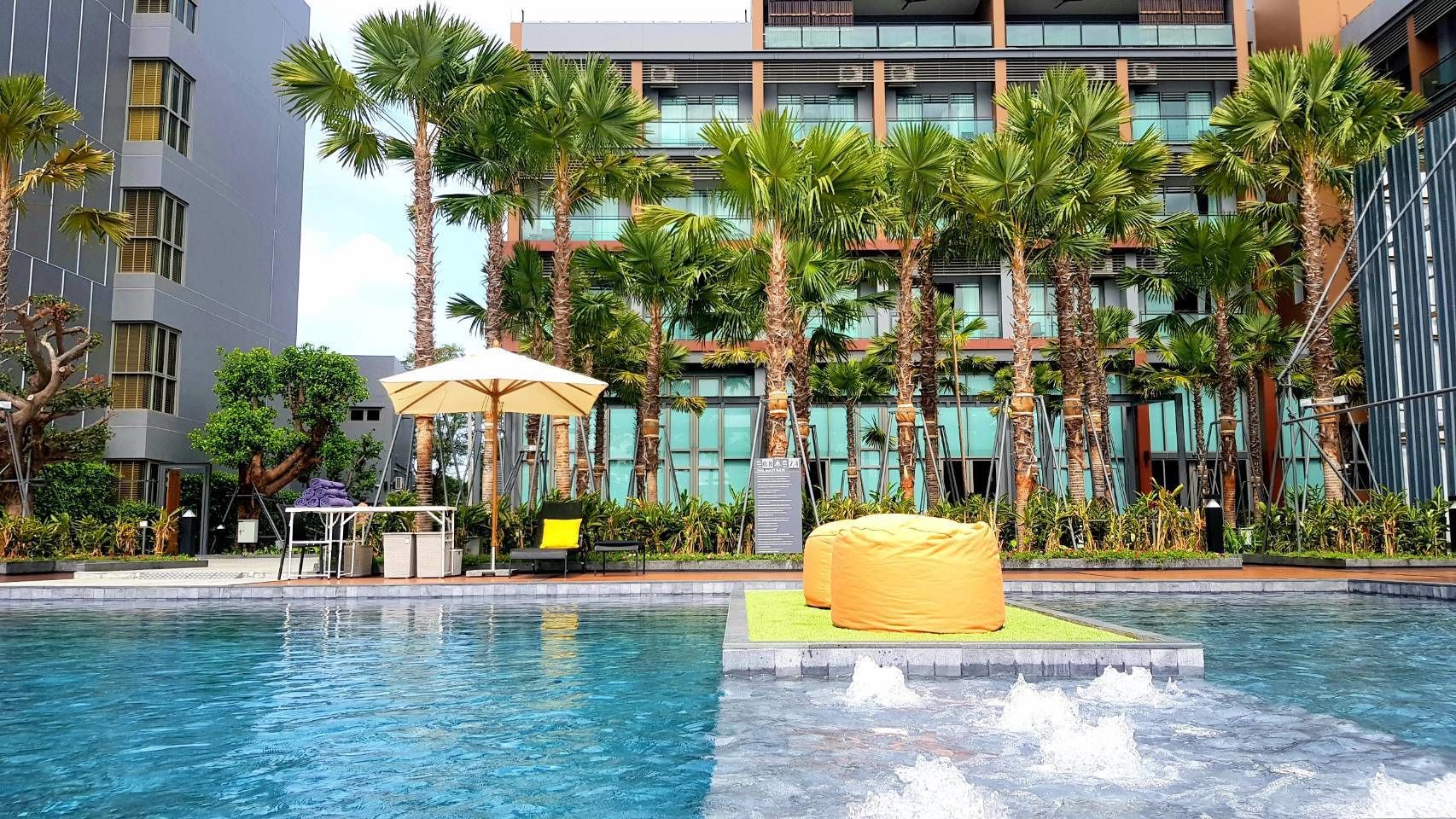 A swimming pool with a water feature, surrounded by palm trees and a modern building with balconies.