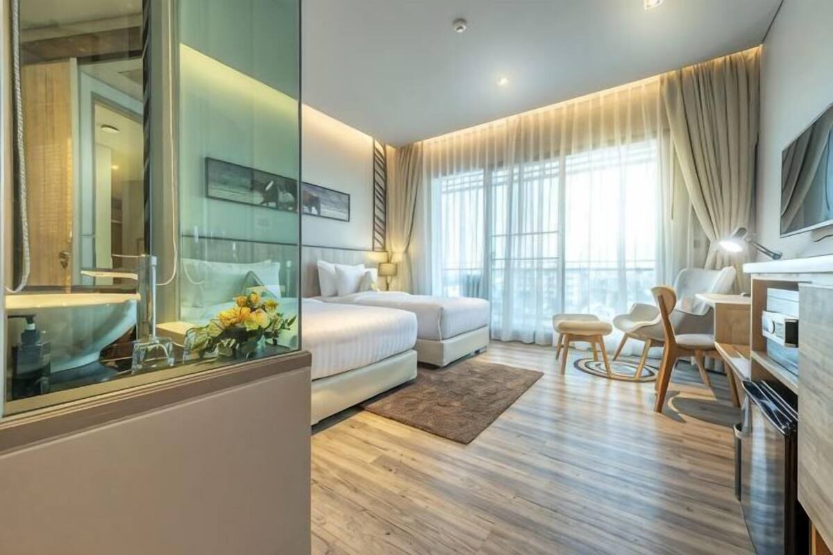 A modern hotel room with a glass partition, a bed, a sofa, a desk, and a TV.