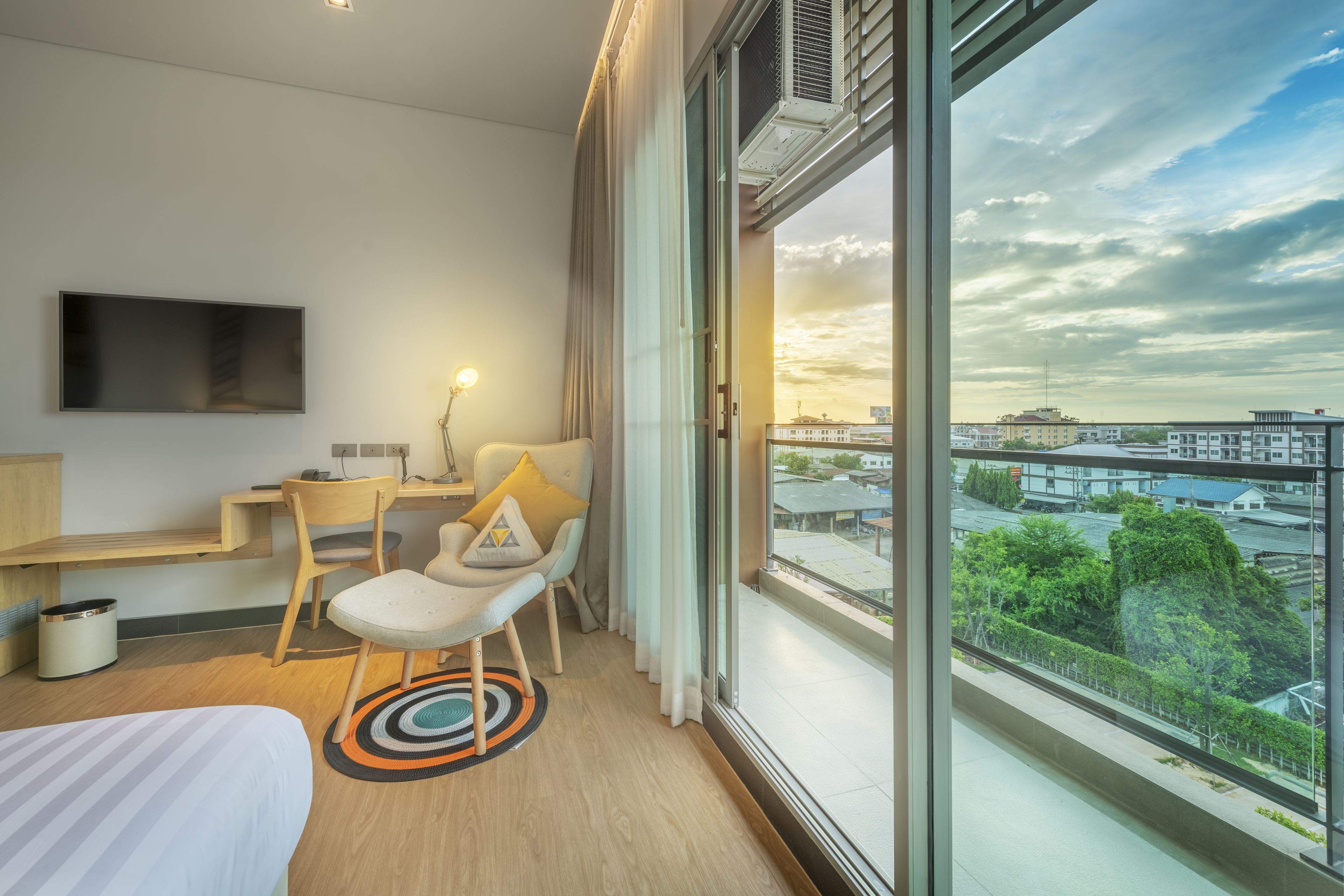 A modern hotel room with a large window, a flat-screen TV, a desk, and a comfortable seating area.