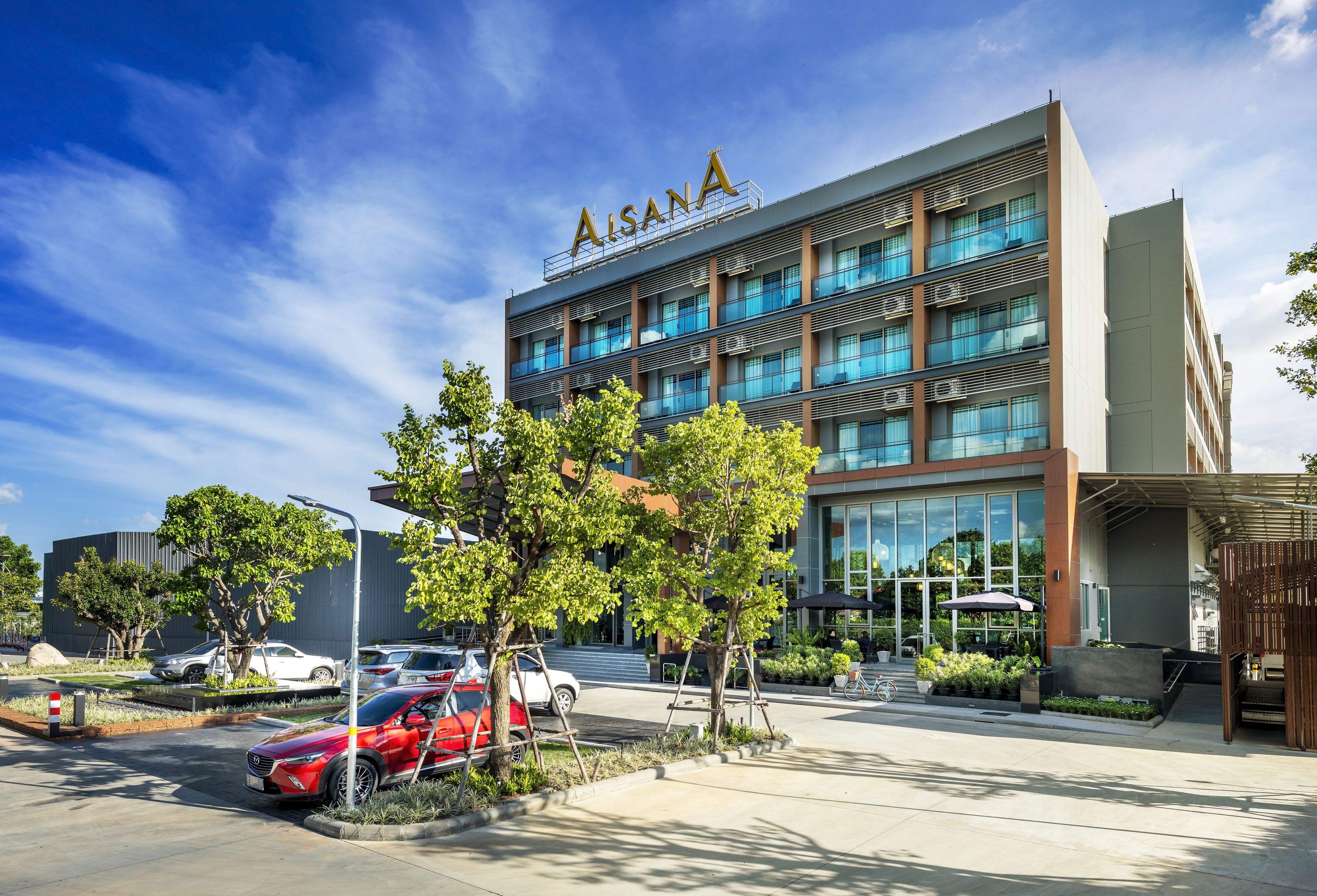 Un edificio moderno con el nombre 'Aisana' en la fachada, que cuenta con varios pisos, ventanas amplias y un área de entrada ajardinada con árboles y un espacio de descanso.