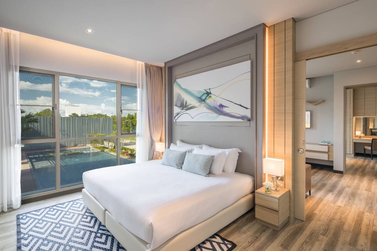 A modern hotel room with a large bed, a painting above the headboard, a view of a swimming pool, and a separate bathroom area.