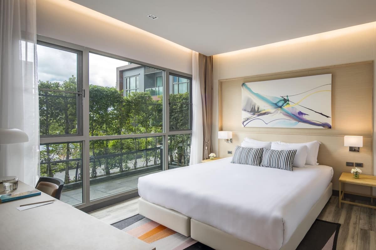 A modern hotel room with a large bed, a desk, and a view of greenery outside.