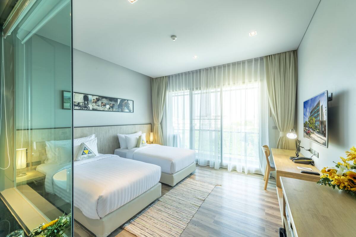 A modern hotel room with two beds, a desk, a chair, a TV, and a large window with curtains.