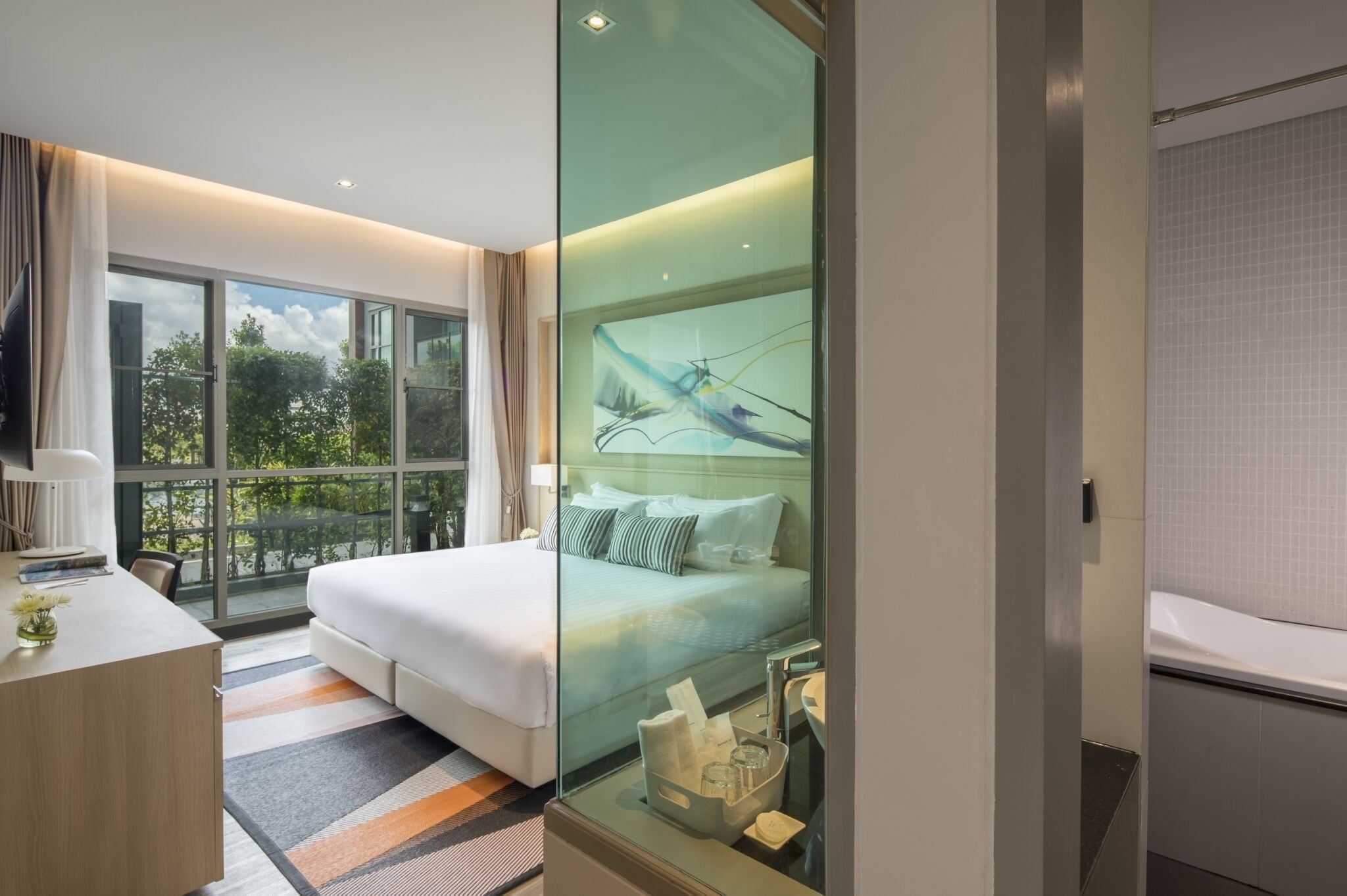 A modern hotel room with a large bed, a desk, and a glass partition.