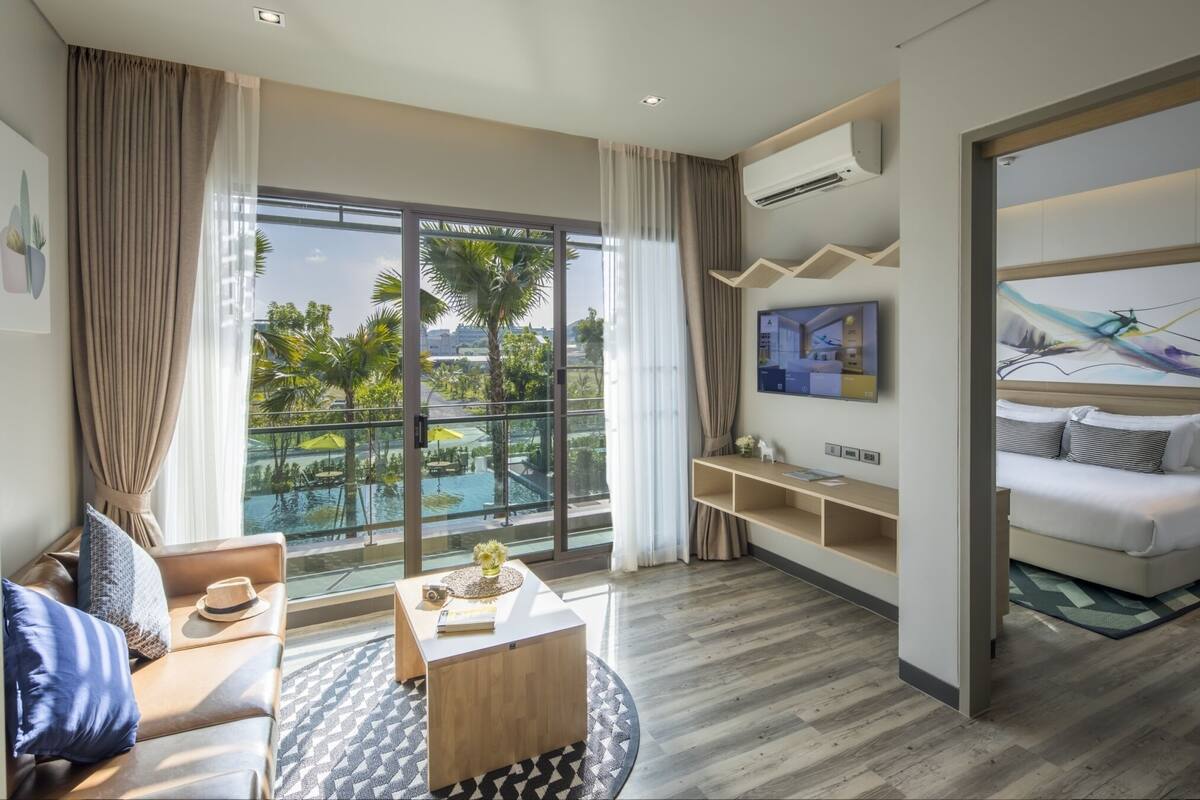 A modern hotel room with a large balcony, a sofa, a coffee table, and a TV mounted on the wall.