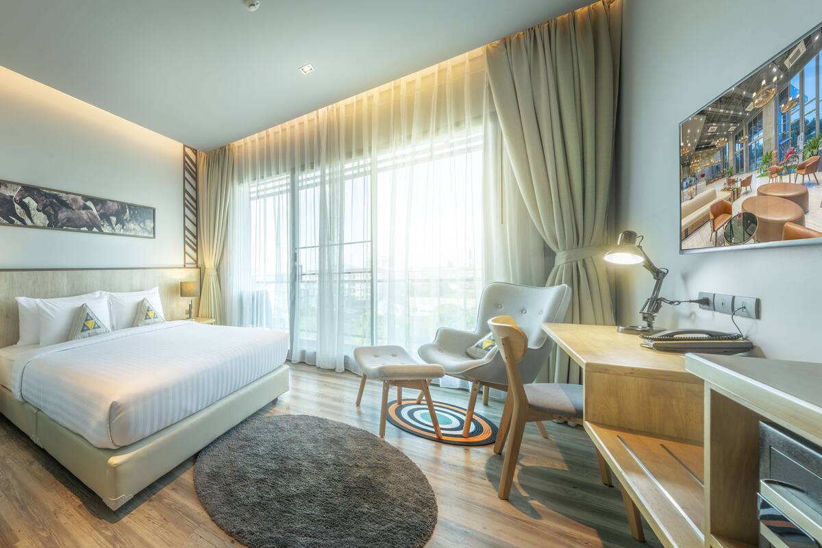 A modern hotel room with a large bed, a desk, a chair, and a view of the city through large windows.