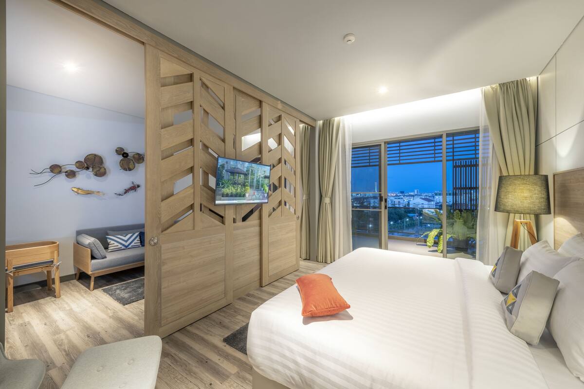 A modern hotel room with a large bed, a TV on a wooden partition, a sofa, and a balcony with a city view.