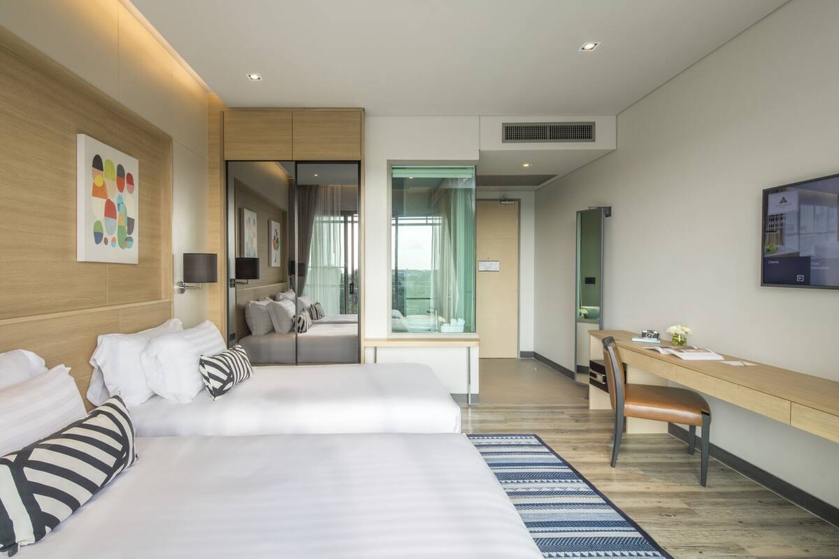 A modern hotel room with two beds, a desk, a chair, and a large window.