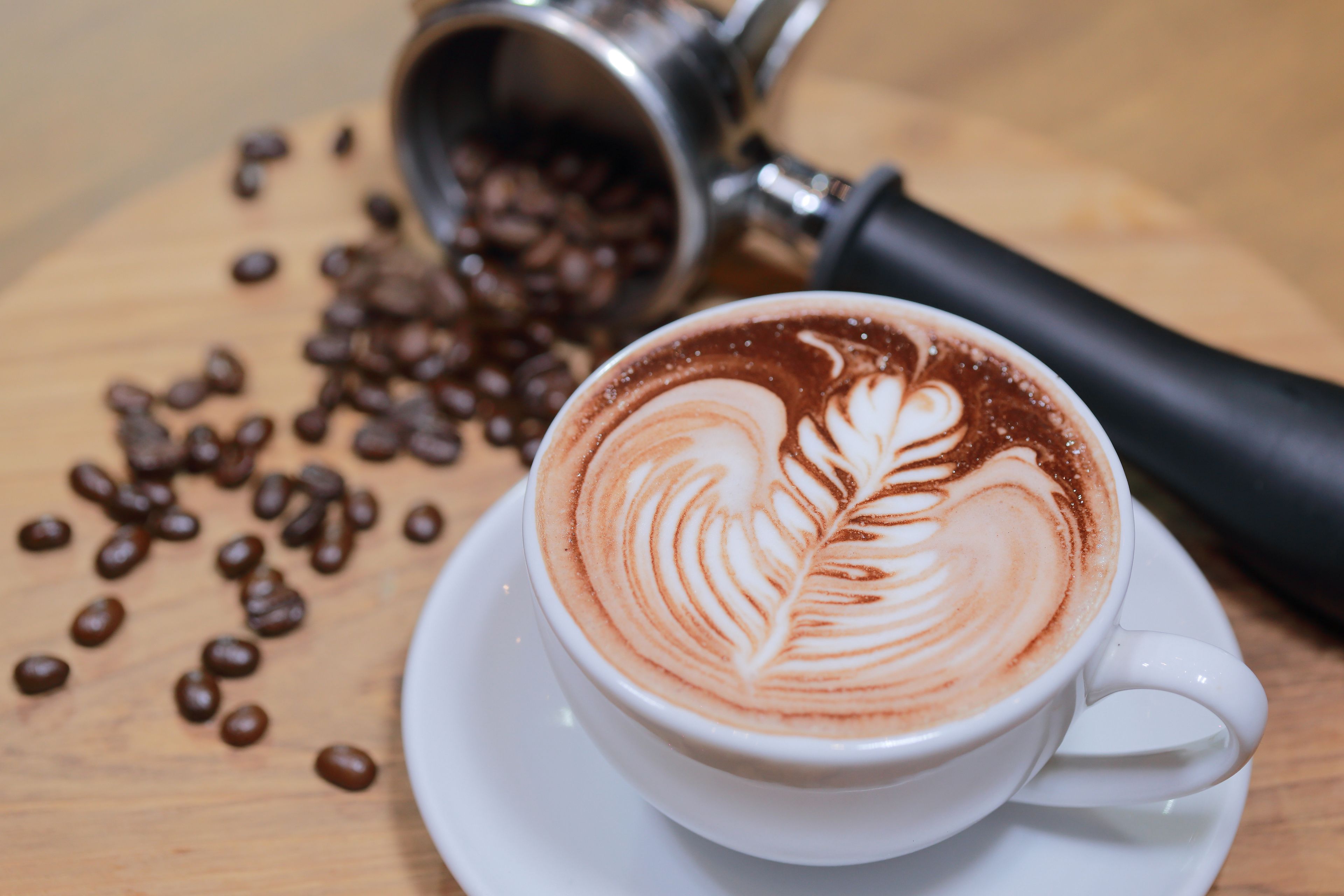 Une tasse de café ornée d’un motif en latte art, posée sur une soucoupe, avec des grains de café éparpillés autour et un moulin à café en arrière-plan.
