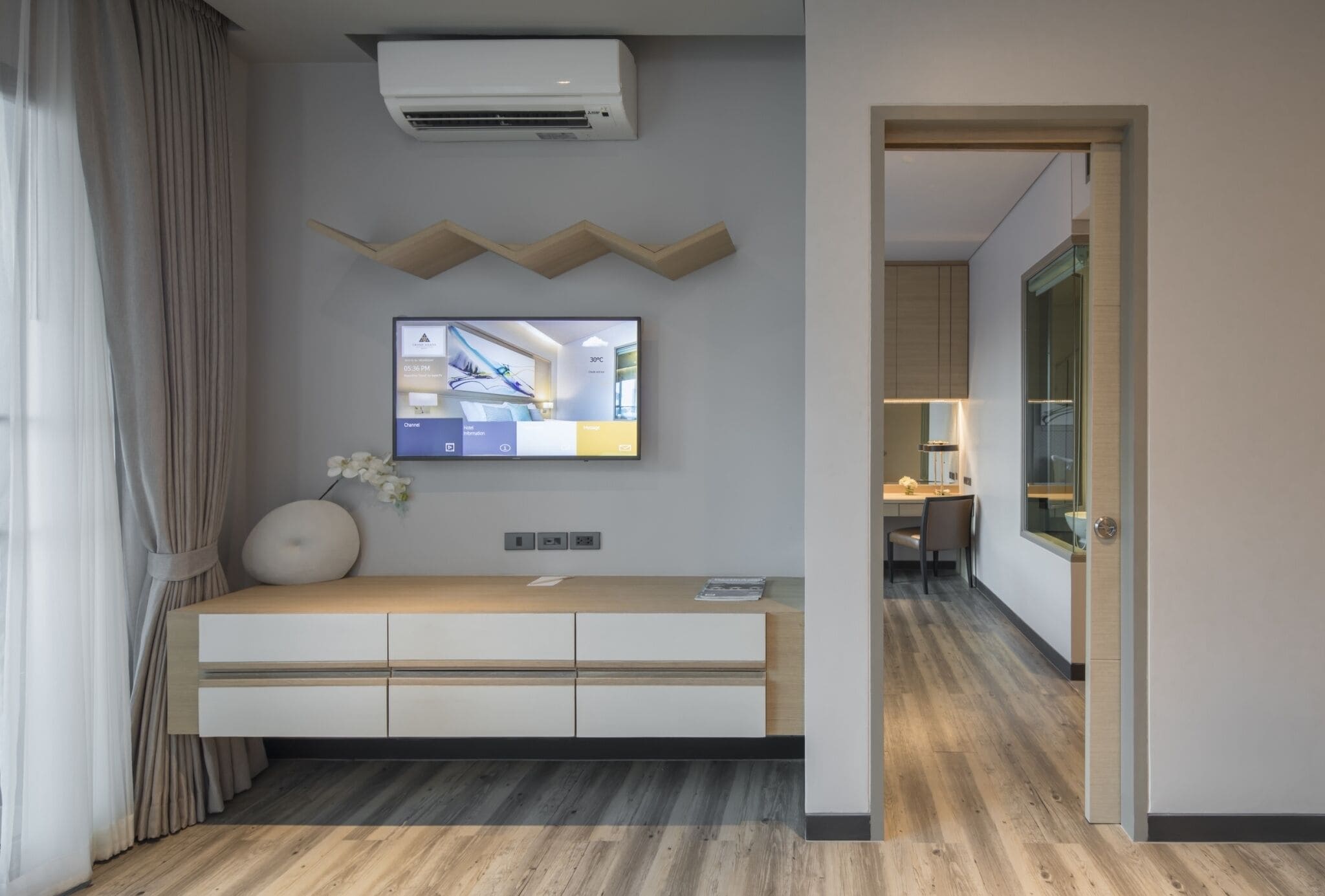 A modern living room with a flat-screen TV mounted on the wall, a wooden console table, and a large window with curtains.