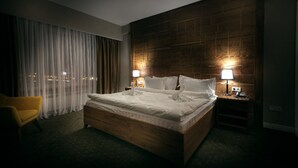 A hotel room with a large bed, two bedside lamps, a wooden headboard, a yellow armchair, and a view of the city at night.