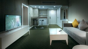 A modern hotel room with a flat-screen TV, a sofa with yellow and gray cushions, a white coffee table, and a kitchenette area.
