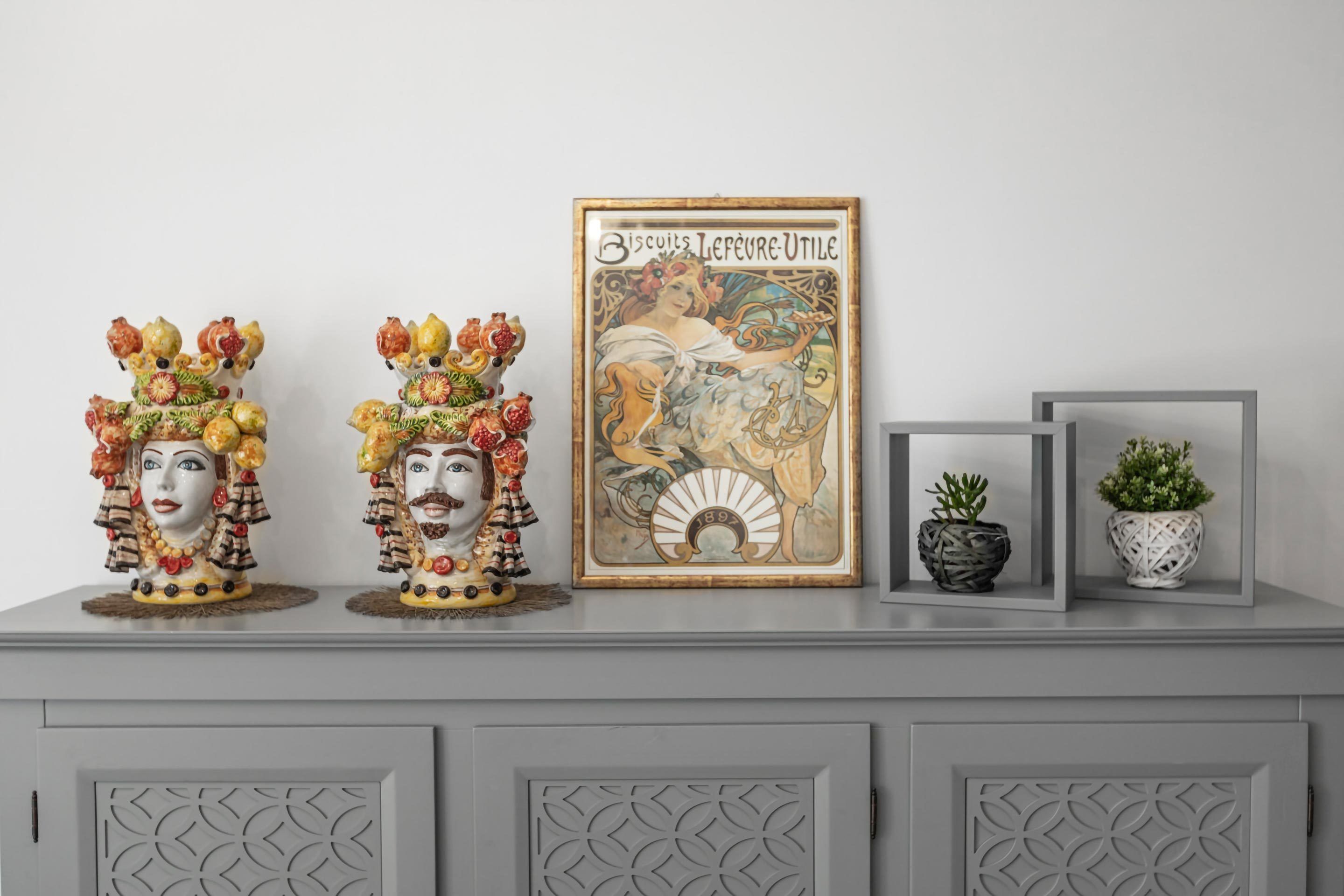 A decorative display with ceramic figures, a framed vintage poster, and potted plants on a grey cabinet.