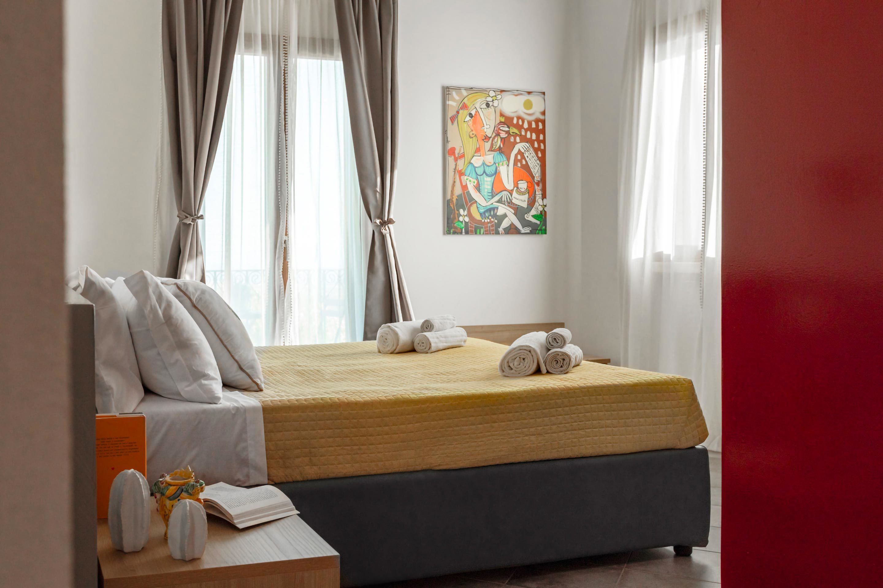 Un lit bien fait, avec des serviettes pliées, une table de chevet sur laquelle se trouvent un livre et un objet décoratif, et un tableau accroché au mur.