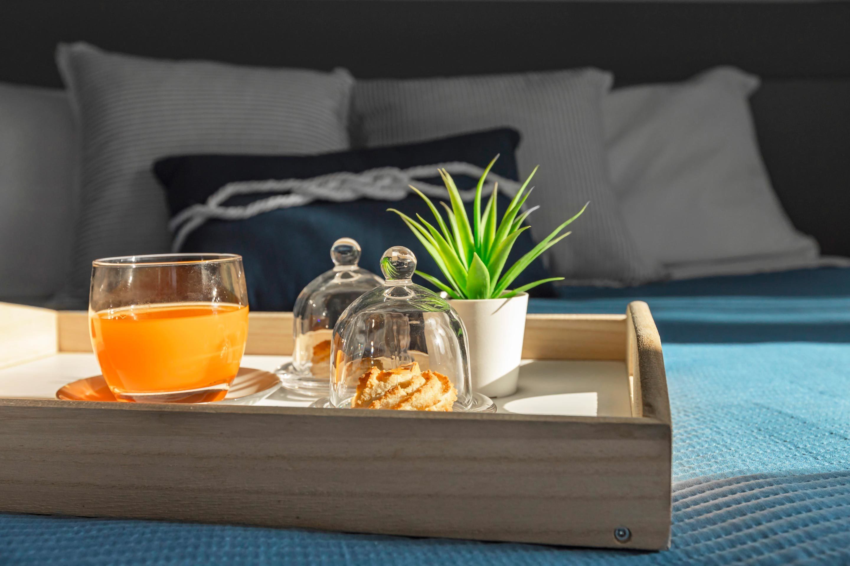 A tray with a glass of orange liquid, a covered container, and a small potted plant on a bed with pillows and a blue blanket.