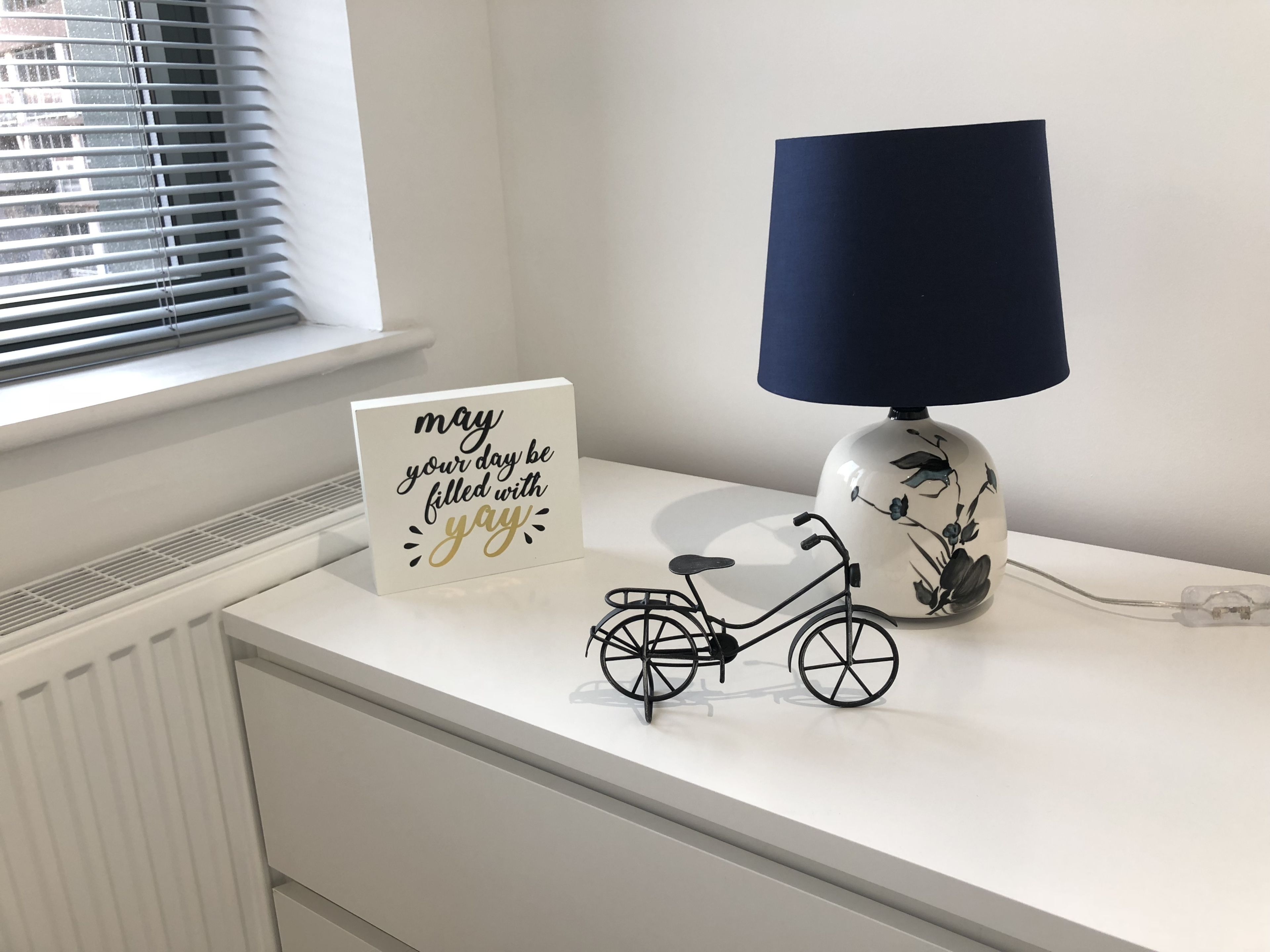 Ein weißer Nachttisch mit einer dekorativen Lampe, einem Schild mit einer positiven Botschaft und einem kleinen Fahrradmodell.