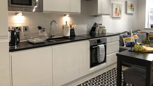 A modern kitchen with white cabinets, a black countertop, stainless steel appliances, and a dining area with a table and chairs. - Cosy One Bedroom Apartment By Creatick (Reading)