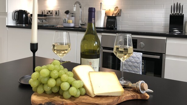 A kitchen counter with a bottle of wine, two glasses of wine, a candle, a bunch of grapes, and a piece of cheese. - Cosy One Bedroom Apartment By Creatick (Reading)